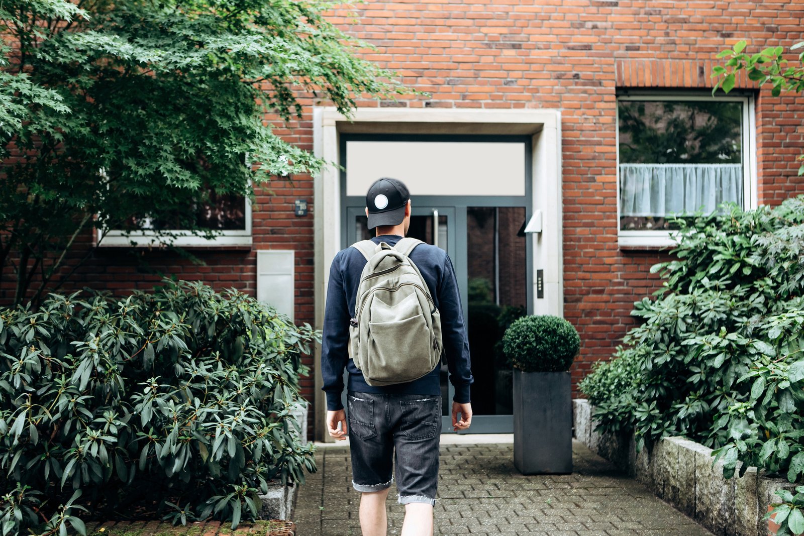 A man wearing a backpack walking toward the front door of a brick house.